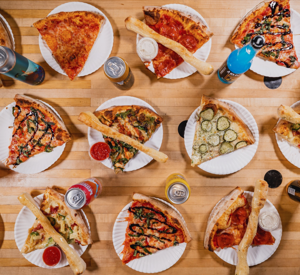 Overhead view of a wooden table covered with various slices of pizza on paper plates, accompanied by breadsticks, dipping sauces, and canned drinks.